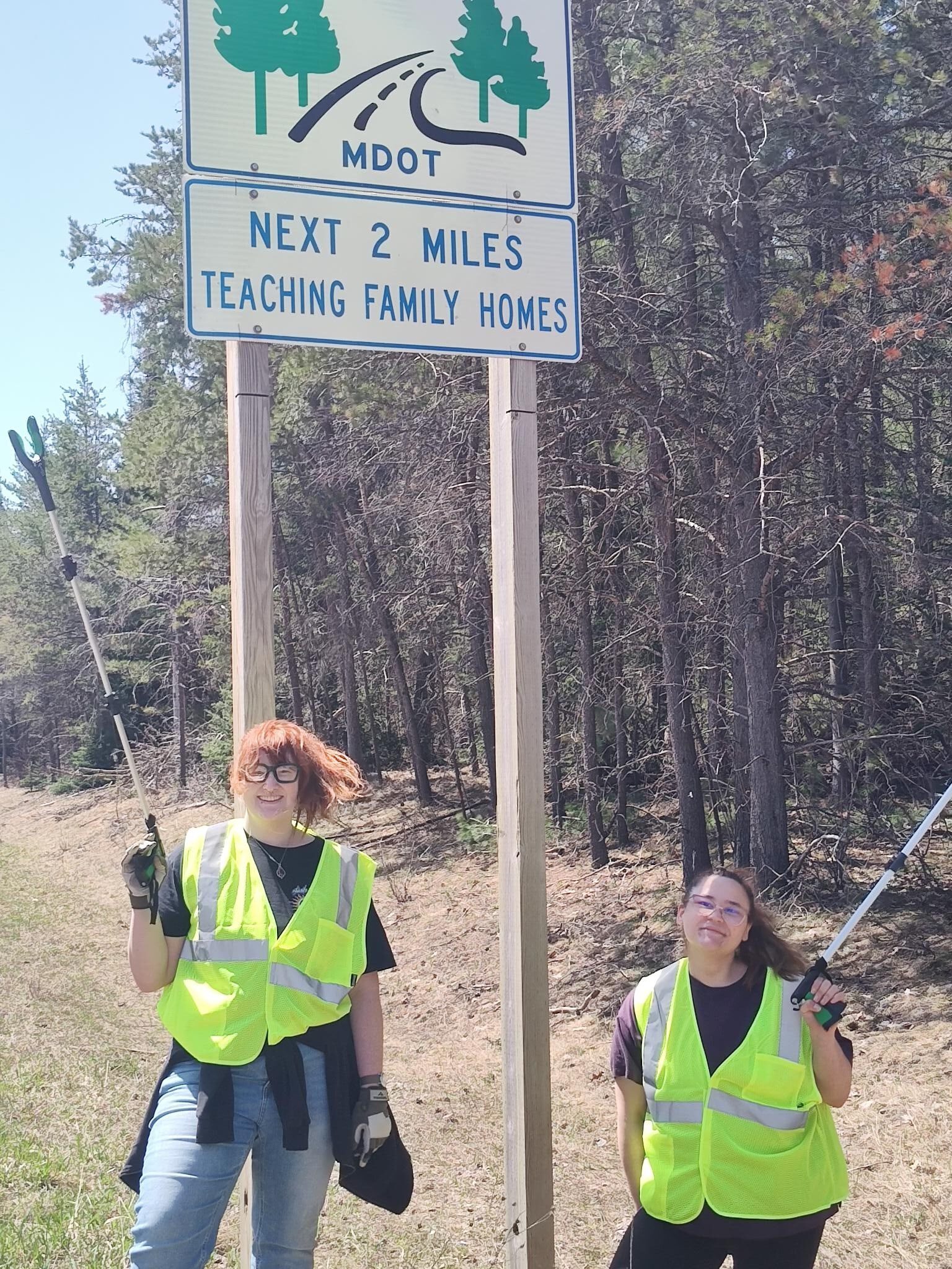 Two volunteers in high-visibility safety vests smiling while standing next to an official "Adopt-A-Highway" sign for Teaching Family Homes.